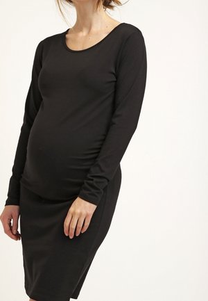 Gravid kvinde iført en tætsiddende sort langærmet kjole, der står med hænderne afslappet ned langs siderne mod en ensfarvet baggrund.