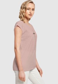 Hellrosa Kurzarm-T-Shirt mit rundem Halsausschnitt und schwarzem Text "ICH BIN ANDERS", der vertikal auf der Vorderseite aufgedruckt ist. Weicher, glatter Stoff.