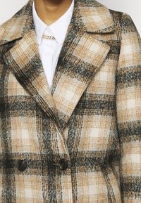 Cappotto a doppio petto a quadri nei toni del marrone, beige e grigio, caratterizzato da una texture in lana, rever a scollatura, e bottoni scuri. Camicia bianca con colletto sotto.