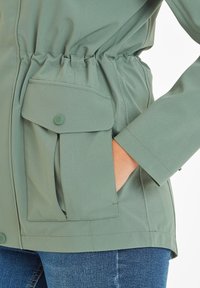 Green, zip-up jacket with elastic waistband; features a large, buttoned patch pocket and smooth, textured fabric; long sleeves.