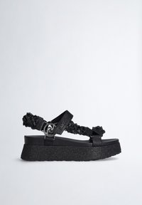 Sandale à plateforme noire avec brides tressées texturées, boucle argentée décorative et semelle épaisse pailletée, présentée sur fond blanc.