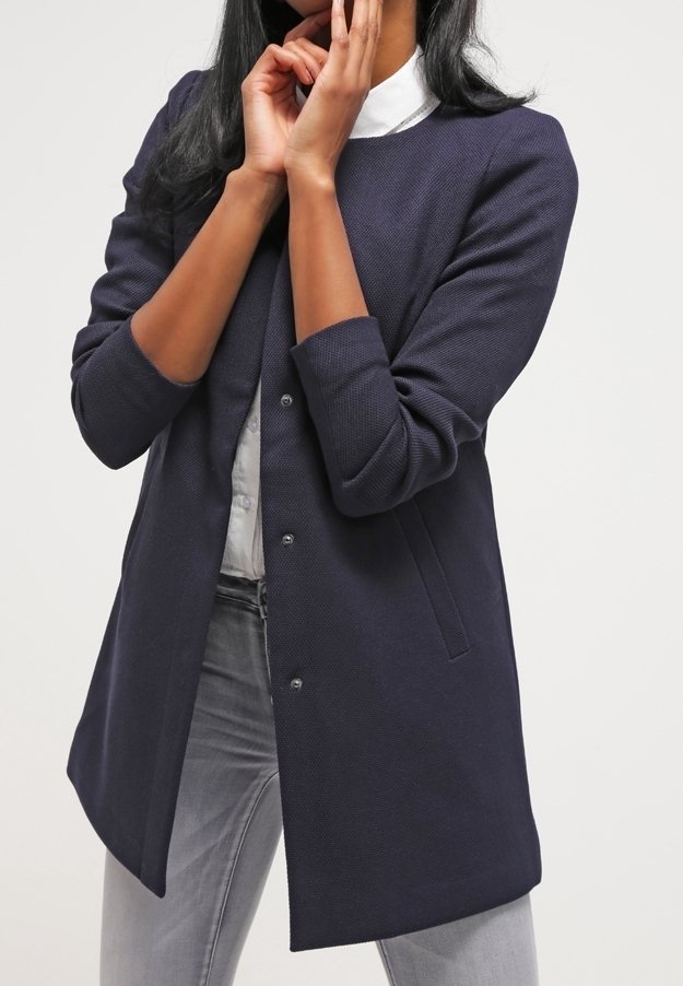 Mörkblå jacka med texturerat tyg, med rund halsringning, tryckknappar och sidofickor, bärs över en ljus knäppskjorta.