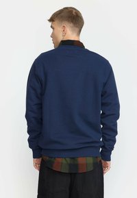 Marineblauwe sweatshirt van katoen. Heeft lange mouwen en een ribbelboord. Gelaagd over een groen en bruin geruit overhemd.