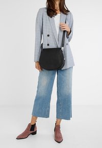 Borsa a tracolla in pelle nera con forma semicircolare, caratterizzata da una texture liscia e accessori in argento, indossata sopra un blazer grigio e jeans.