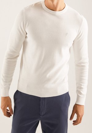 Weißer Strickpullover mit rundem Ausschnitt, gerippten Bündchen und Saum, mit dezent gesticktem Logo auf der Brust. Kombiniert mit dunkelblauen Hosen.