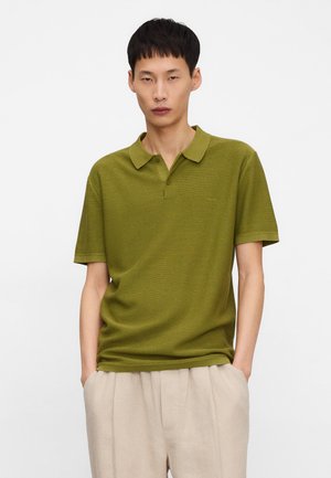 Młody mężczyzna w oliwkowozielonej koszulce polo z krótkim rękawem i beżowych spodniach, z rękami w kieszeniach, stoi na tle jednolitego białego tła.