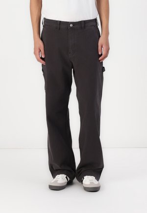 Mężczyzna noszący luźne czarne spodnie typu cargo i białe trampki, stojący na tle jednolitego białego tła.