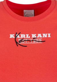 Punane puuvillane T-särk, millel on ümar kaelus, valge tekst "KARL KANI" ja musta värvi tikand.