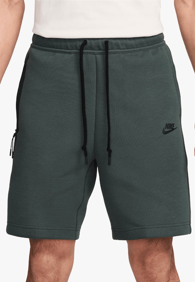 Gröna sportshorts tillverkade av en blandning av bomull, med svarta draremmar, sidofickor och en liten svart Nike-logotyp på nedre vänstra sidan.