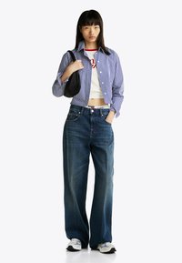 Camisa de botones a rayas en azul y blanco, camiseta blanca corta con estampado rojo, jeans de mezclilla oscuros de pierna ancha y zapatillas. Bolsa de mano negra.