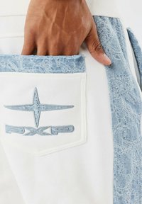 Witte stof met lichtblauwe denimaccenten op de zak en zijkant. Geborduurd logo in het blauw op de zak. Zachte textuur met rafelige randen.