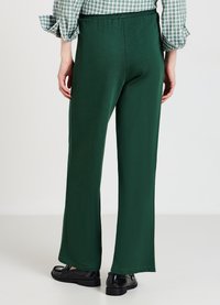 Person mit dunkelgrünen weit geschnittenen Hosen, schwarzen glänzenden Slippern und einem grün-weiß karierten Hemd, steht vor einem schlichten weißen Hintergrund.