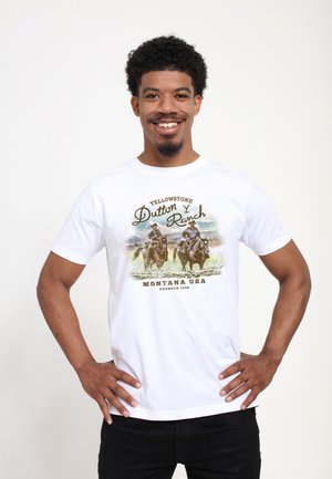 Hombre sonriente con pelo rizado está de pie con las manos en las caderas, llevando una camiseta blanca con dos jinetes a caballo y el texto "Yellowstone Dutton Ranch Montana USA".
