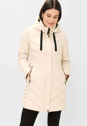 Woman wearing a cream quilted hooded winter coat with black zipper pockets and black drawstrings, hands in pockets, looking down and smiling.