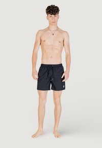 I pantaloni da bagno neri presentano un cinturino elastico con cordino e un accento con logo. Il tessuto sembra leggero e a asciugatura rapida.