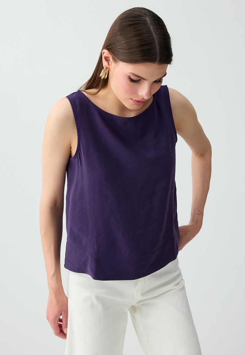Femme aux cheveux bruns et raides, portant des boucles d'oreilles en or, un haut violet foncé sans manches et un pantalon blanc, regardant vers le bas avec la main sur la hanche.
