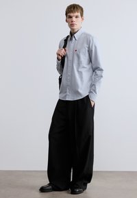 Φόρεμα με ανοιχτό μπλε πουκάμισο με τσέπη στο στήθος, μαύρο παντελόνι με φαρδιά μπατζάκια και μαύρα παπούτσια. Το μοντέλο ποζάρει με μια τσάντα ώμου.