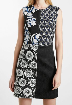 Robe sans manches avec quatre panneaux à motifs noirs et blancs comprenant des dessins floraux, abstraits et en éventail, portée par une personne debout.