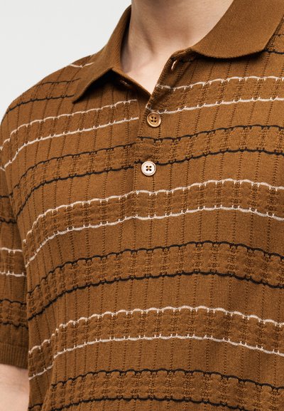 Κοντινό πλάνο ενός καφέ πλεκτού πόλο πουκαμίσου με υφή οριζόντιων ραβδώσεων και δύο ταν κουμπιά στη λαιμόκοψη.