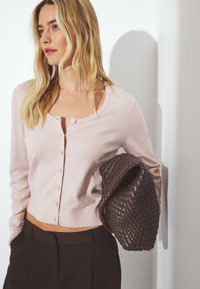 Gaiši rozā kardigāns ar apaļu kakla izgriezumu un pogu aizdari, kombinēts ar tumšu austu rokassomu. Gluda tekstūra un pieguļošs dizains.