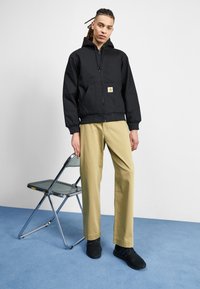 Black hooded jacket with front zipper and logo patch, paired with beige pants and black sneakers, standing beside a metal chair on blue floor.