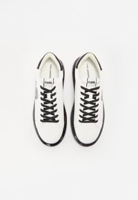 Białe trampki z czarnymi sznurowadłami i czarnymi błyszczącymi akcentami. Gładka skórzana cholewka z zaokrąglonym noskiem i detalem z logo po boku.