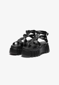 Black platform sandals with crisscrossed straps, faux leather material, textured sole, and adjustable buckles for a secure fit.
