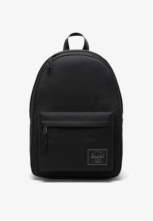 Mochila de tela negra con forma redondeada, bolsillo frontal con cremallera y un parche de logo. Cuenta con correas ajustables y un asa superior.