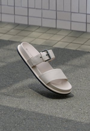 Sandale beige à bout ouvert avec deux larges brides, dont une avec une boucle en métal, sur un sol en pavé gris texturé près d'un mur carrelé.