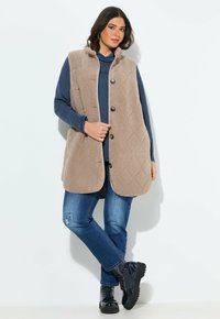 Donna che indossa un gilet in pile beige trapuntato sopra un hoodie blu, abbinato a jeans blu e stivali neri con lacci, in piedi contro uno sfondo bianco.