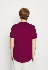 Persona con cabello rojo corto, vistiendo una camiseta magenta sencilla y pantalones cortos beiges, vista desde atrás contra un fondo blanco.