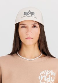 Beige caps med et frontlogo, har en strukturert design og mesh-paneler. Modellen har på seg en lysebrun sweatshirt med hvit tekst.