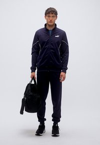 Σκούρο μπλε φόρμα από ελαφρύ ύφασμα με λευκές λεπτομέρειες, ζακέτα με φερμουάρ, ελαστικά μανσέτες και παντελόνι που ταιριάζει. Μαύρο σακίδιο πλάτης στο χέρι.