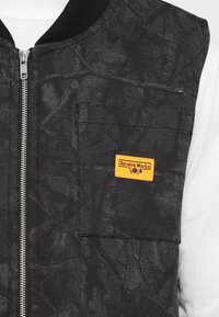 Μαύρο γιλέκο bomber με υφασμάτινη υφή και μοτίβο. Διαθέτει εμπρόσθιο φερμουάρ και μια μικρή πορτοκαλί ετικέτα «Service Works» στην τσέπη του στήθους.