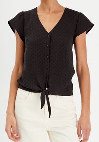 Blouse noire à manches courtes avec un col en V, une fermeture à boutons, un tissu texturé et un détail de ourlet noué. Associée à un pantalon de couleur claire.