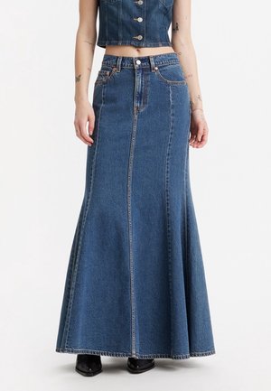 Femme portant une jupe longue en jean bleu évasée taille haute, un crop top en jean boutonné et des chaussures noires, debout devant un fond uni.