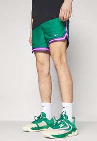 Pantalones cortos verdes de Nike con ribetes morados y detalles laterales. Combinados con zapatillas verdes que presentan un logo rosa, calcetines blancos y una camiseta negra.