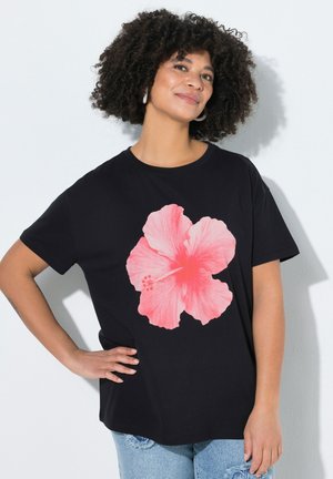 Kobieta o kręconych włosach, ubraną w czarny t-shirt z dużym różowym nadrukiem kwiatu hibiskusa, stojąca z jedną ręką na biodrze.