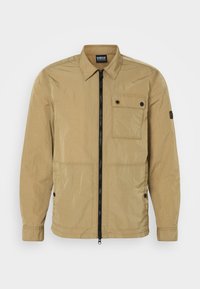 Veste beige légère au texture froissée avec une fermeture éclair sur le devant, poche poitrine boutonnée et poches latérales. Présente un design avec col.