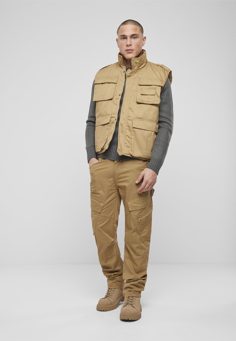 Homme debout, les mains partiellement dans les poches, portant un pantalon cargo beige, un gilet assorti avec plusieurs poches, un pull gris à manches longues et des bottes beige.