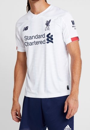 Uomo che indossa la maglia da calcio bianca del Liverpool FC con il logo Standard Chartered e pantaloncini Adidas blu navy su sfondo semplice.