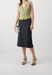 Grön ärmlös väst med djup V-ringning och tre knappar, kombinerad med en knälång svart kjol och svarta klackar med remmar, håller en liten väska.