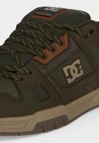 Κοντινή λήψη ενός πράσινου και καφέ sneaker της μάρκας DC με κορδόνια με σχέδιο και μπεζ λαστιχένια σόλα.