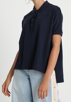 Femme portant un polo ample bleu marine à manches courtes et un jean bleu clair avec des panneaux latéraux blancs et des boutons-pression.