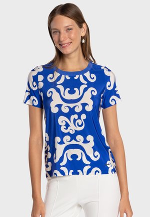 Top azul de manga corta con patrón blanco en espiral, escote redondo y textura suave. Combinado con pantalones blancos, modelo sonriendo.