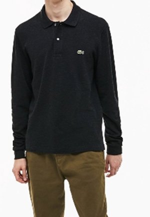 Schwarz longsleeve Poloshirt mit Kragen und einem dezenten grünen Logo auf der Brust, getragen zu hellbraunen Hosen. Weicher Stoff.