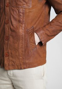 Person klädd i en brun läderjacka med knappar, handen nerstoppad i sidofickan, tillsammans med ljusbeiga byxor.