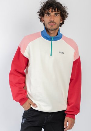 Hombre con cabello rizado que lleva un jersey de bloques de color con cuello azul, parte delantera crema, hombros rosas y mangas rojas, de pie frente a un fondo blanco liso.