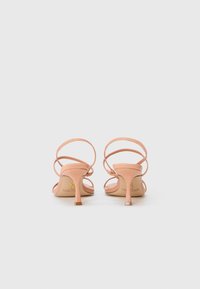 Ett par nude rosa högklackade sandaler med tunna ankelremmar, visade bakifrån på en enkel vit bakgrund.