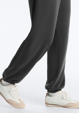 Gambe con pantaloni della tuta grigio scuro e sneakers bianche con dettagli beige su sfondo bianco.